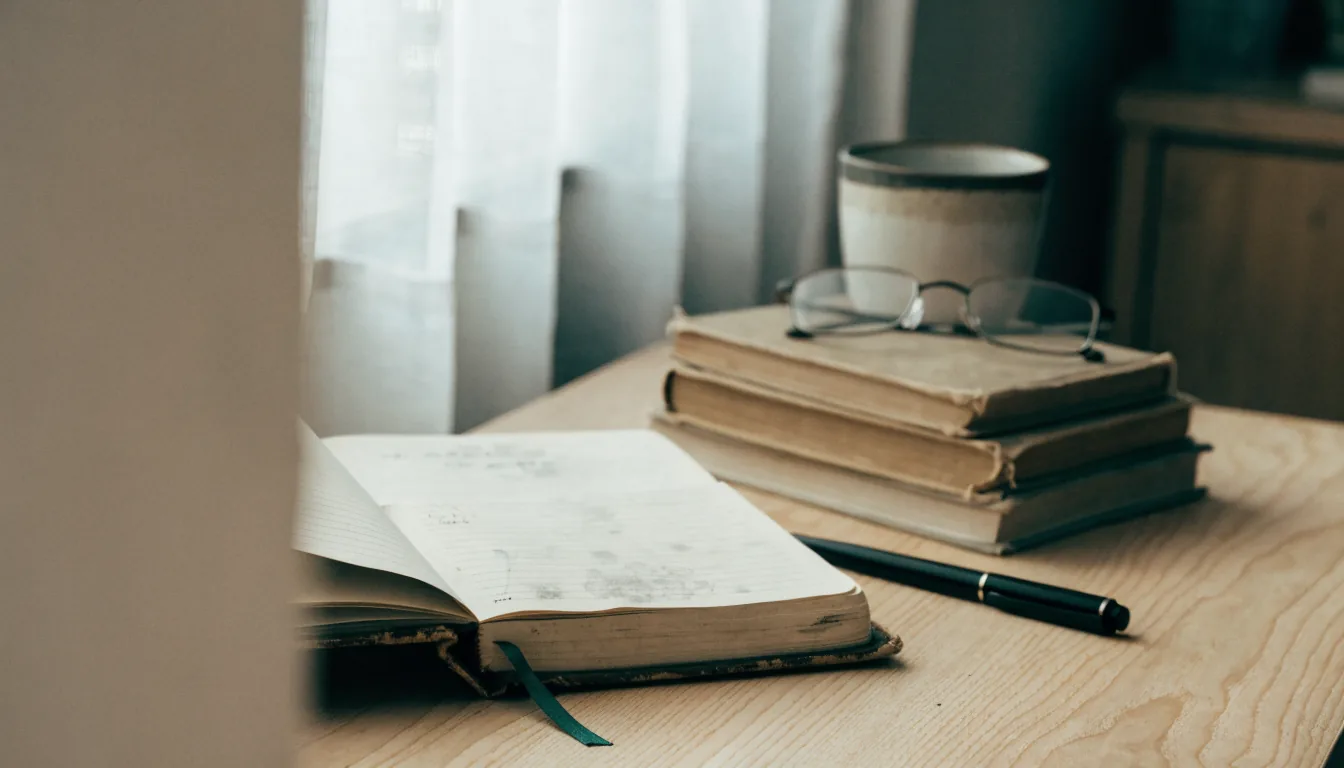 A quiet personal workspace: a worn leather-cornered notebook open beside a fountain pen, a small stack of reference books, a mug and folded reading glasses.