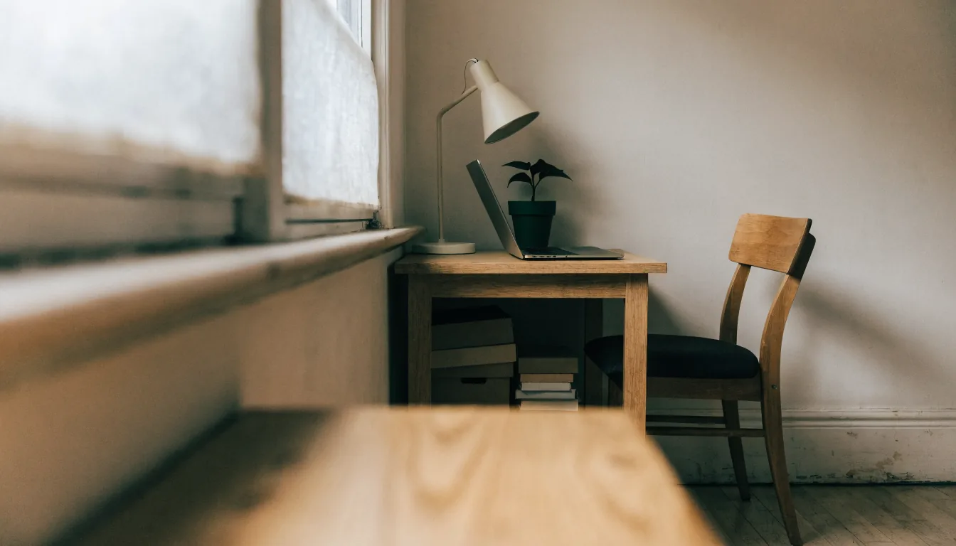 A home office corner in a spare room: a small wooden desk against a plain wall with a laptop, lamp, plant and a low shelf of box files, domestic skirting board visible.