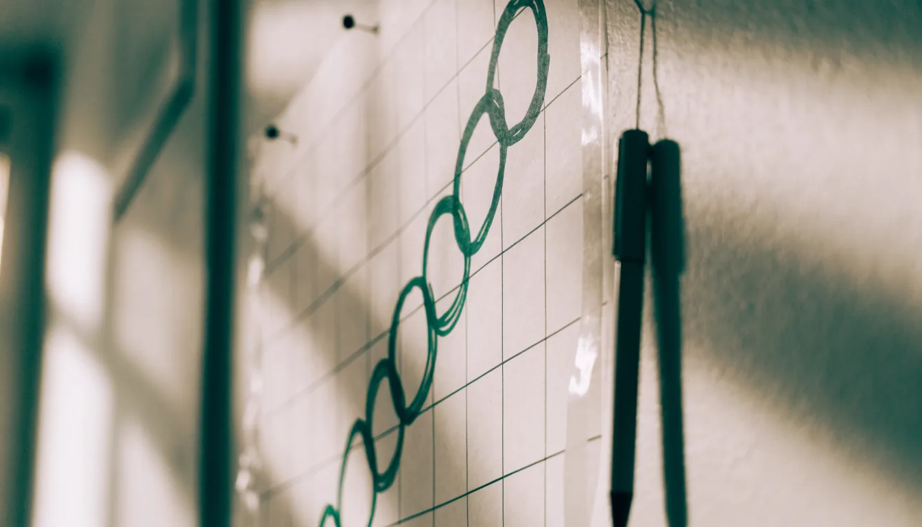 A blank paper grid chart pinned to a wall with green marker circles drawn around squares at regular repeating intervals, and a pen hanging beside it on a string.